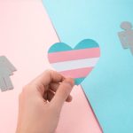 Hand holding a paper heart with transgender pride flag. Paper figures of woman and man on pink and blue background. Trans people and gender identity.