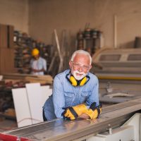 Mature elderly joiner looking is leaned on a workbench at the carpentry workshop. He is looking at the camera and smiling